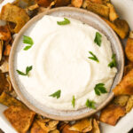 A bowl of creamy white toum garnished with parsley, surrounded by pieces of toasted pita chips.
