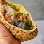 A hand holding a grilled sabich sandwich filled with roasted eggplant, chopped tomatoes, cucumbers, herbs, and a creamy white sauce.