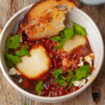 A bowl of vegan shakshuka topped with vegan feta cheese, fresh parsley, and served with a piece of toasted bread.