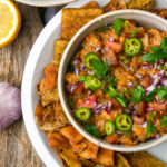 Bowl of chunky ful medames garnished with sliced jalapeños, red onion, parsley, and tomatoes, surrounded by pita chips on a white plate. Lemon and garlic are nearby on a wooden surface.