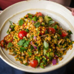 A plate of pearl couscous salad with cherry tomatoes, red onions, cucumber, parsley, mint, and chickpeas, garnished with herbs.