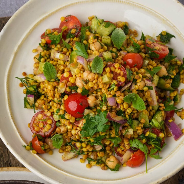 A bowl of pearl couscous salad with cherry tomatoes, chickpeas, chopped cucumber, red onion, fresh herbs, and a mix of grains on a white plate.