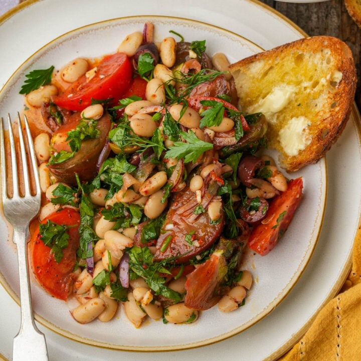 A plate of piyaz salad with tomatoes, red onion, parsley, and olives, served with a slice of buttered toasted bread and a fork on the side.