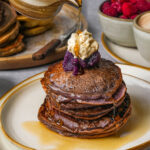 A stack of ube pancakes topped with whipped cream and ube jamis being drizzled with syrup, with bowls of raspberries and sauce in the background.