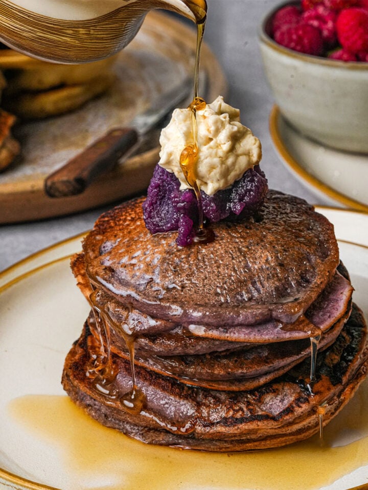 A stack of ube pancakes topped with whipped cream and ube jamis being drizzled with syrup, with bowls of raspberries and sauce in the background.