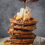 A stack of vegan banana pancakes topped with whipped cream, walnuts, and banana slices, with syrup being poured over them. A fork rests on the plate.