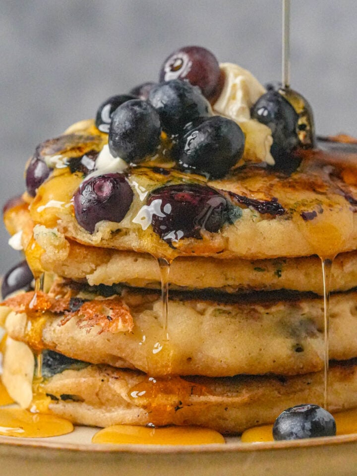 A stack of vegan blueberry pancakes topped with butter, fresh blueberries, and syrup being poured over them, on a beige plate.