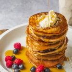 A stack of vegan pancakes topped with butter and syrup, served on a white plate with raspberries and blueberries on the side.