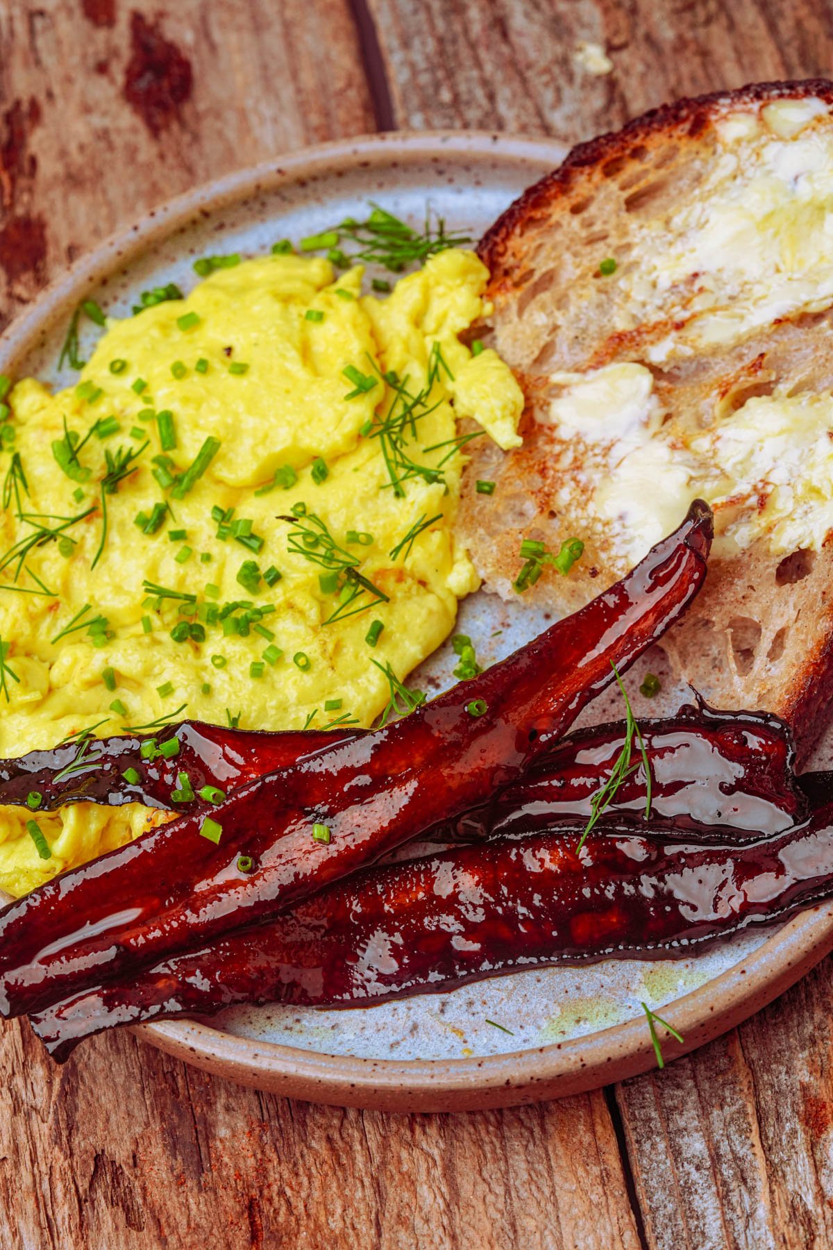 A plate with tofu scrambled eggs topped with chopped herbs, two pieces of glazed carrot bacon, and a slice of buttered toast on a wooden surface.