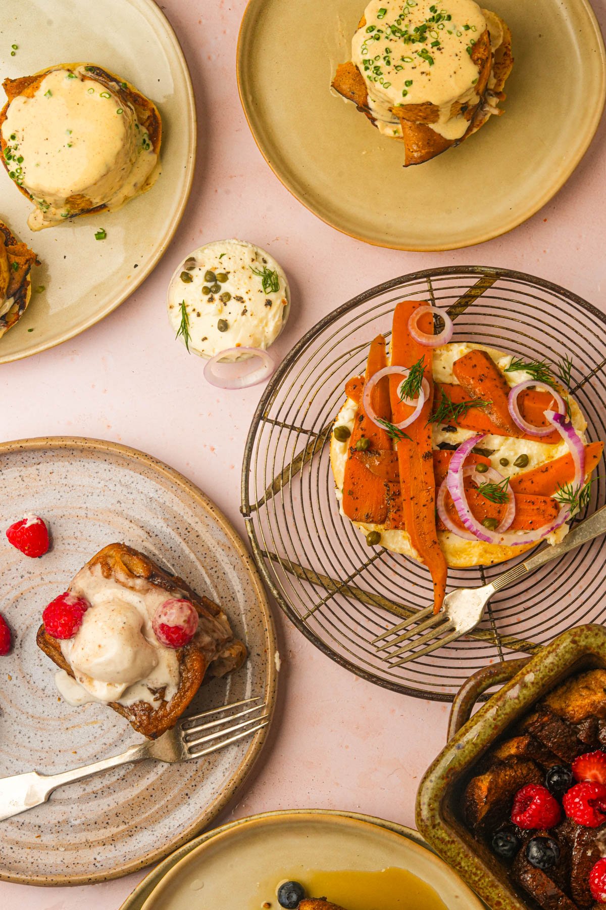 A top-down view of assorted plated brunch dishes, including carrot lox on a bagel, a vegan eggs benedict, Hong Kong French toast with berries, and a baked casserole on a pink surface.