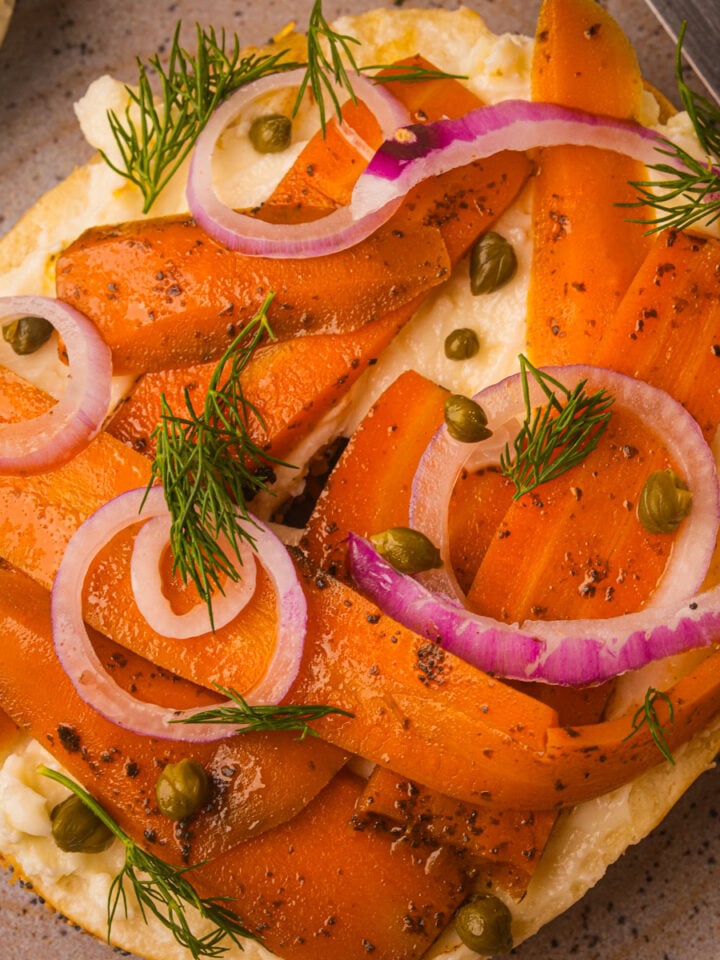A plate with a bagel topped with cream cheese, carrot lox, red onion rings, capers, and fresh dill.