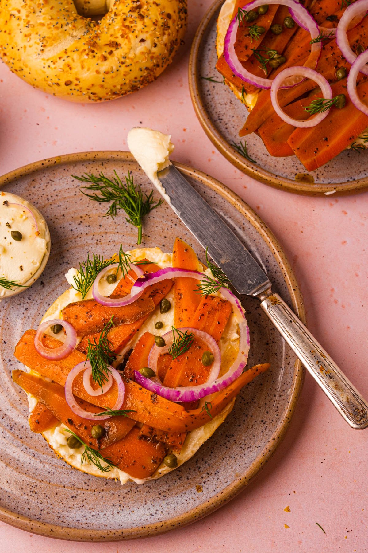 A bagel with cream cheese, carrot lox, red onion rings, capers, and dill on a plate, with a spreading knife and another bagel in the background.