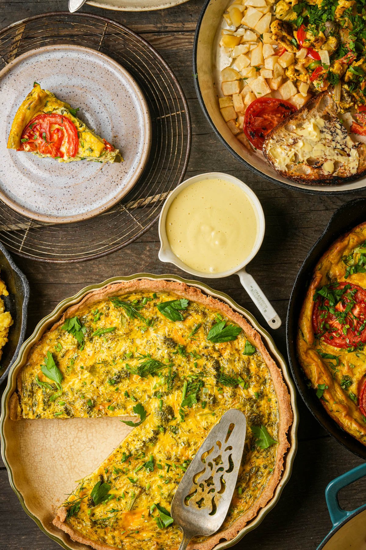 A table set with a vegan quiche, a slice on a plate, a cast iron skillet with a vegan scrambled eggs, a bowl of creamy Just Egg liquid, a vegan quiche, and a vegan frittata, all on a wooden surface.