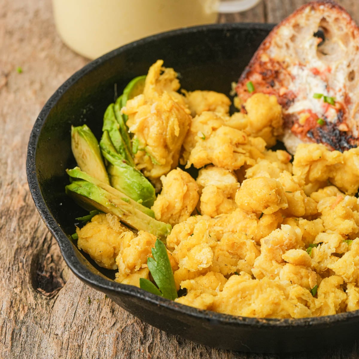 A cast iron skillet filled with scrambled Just Egg, sliced avocado, and a piece of toasted bread on a wooden surface.