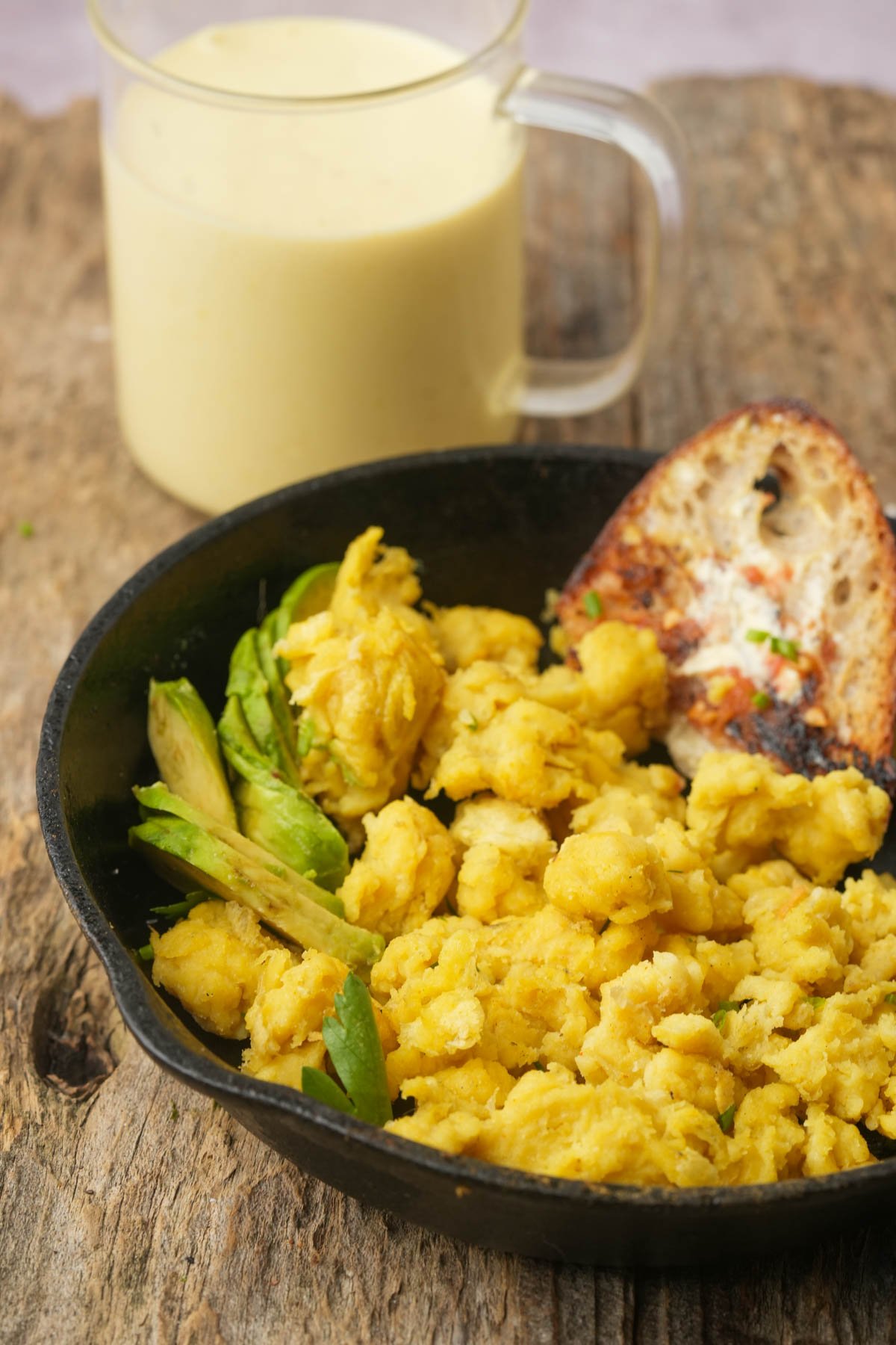 A black pan with vegan scrambled eggs, green garnish, and a slice of toasted bread, placed on a wooden surface; a glass mug of a yellow Just Egg is in the background.