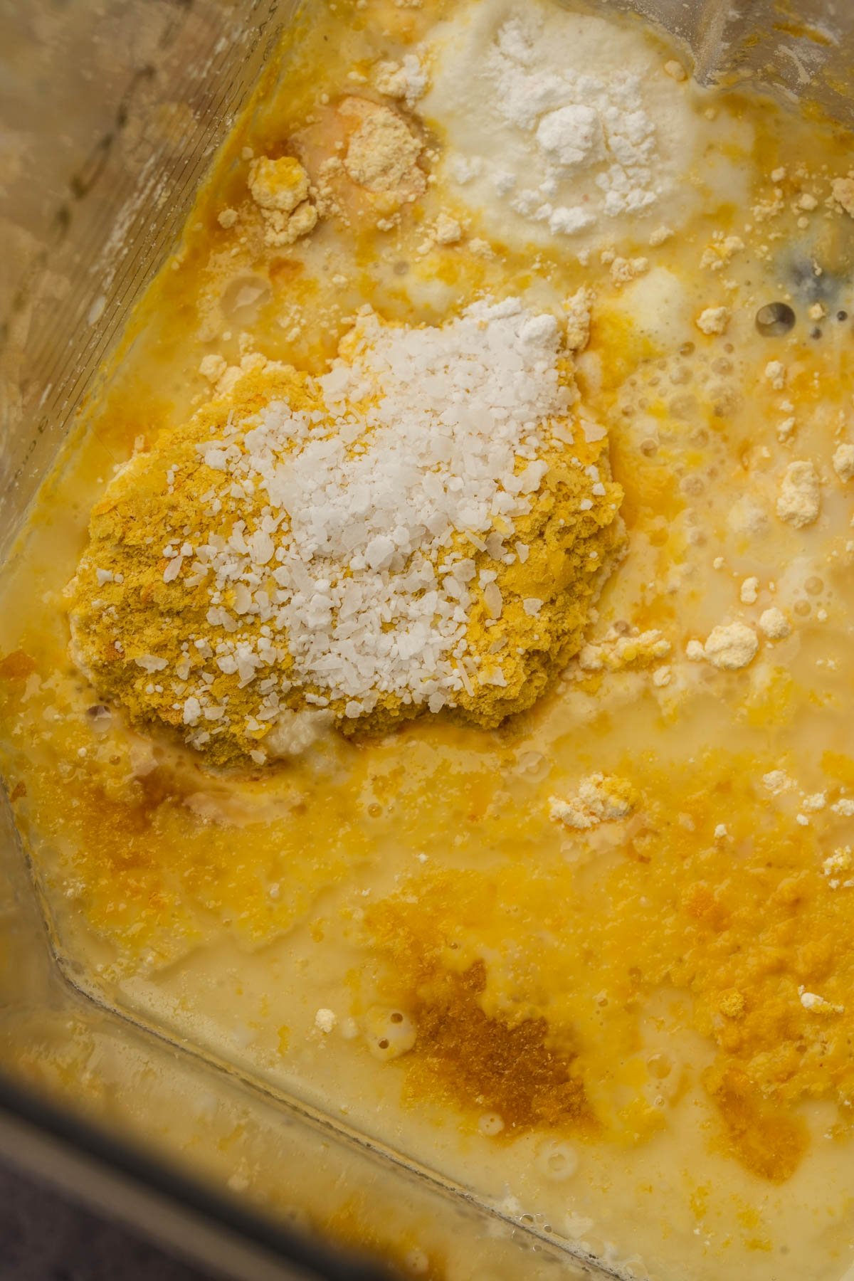 A close-up view of various powdered and liquid ingredients for making Just Egg, including yellow and white powders, in a blender container, ready to be mixed.