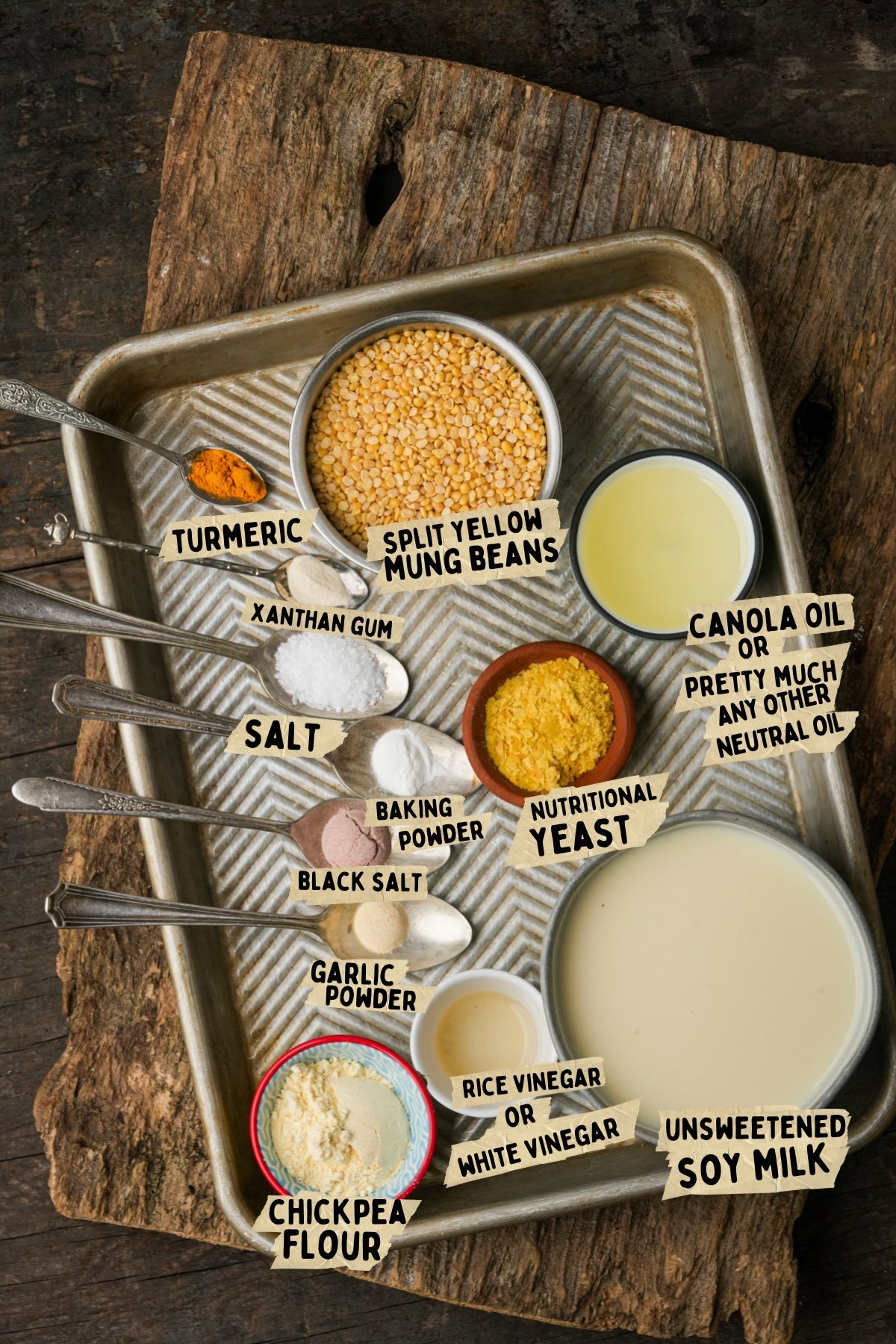 A tray holds labeled ingredients in small bowls and spoons, including split yellow mung beans, turmeric, chickpea flour, nutritional yeast, oils, soy milk, and various powders.