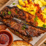 Plate with crispy bacon strips, scrambled eggs topped with chili flakes and herbs, a small dish of red sauce, and a slice of toasted bread.