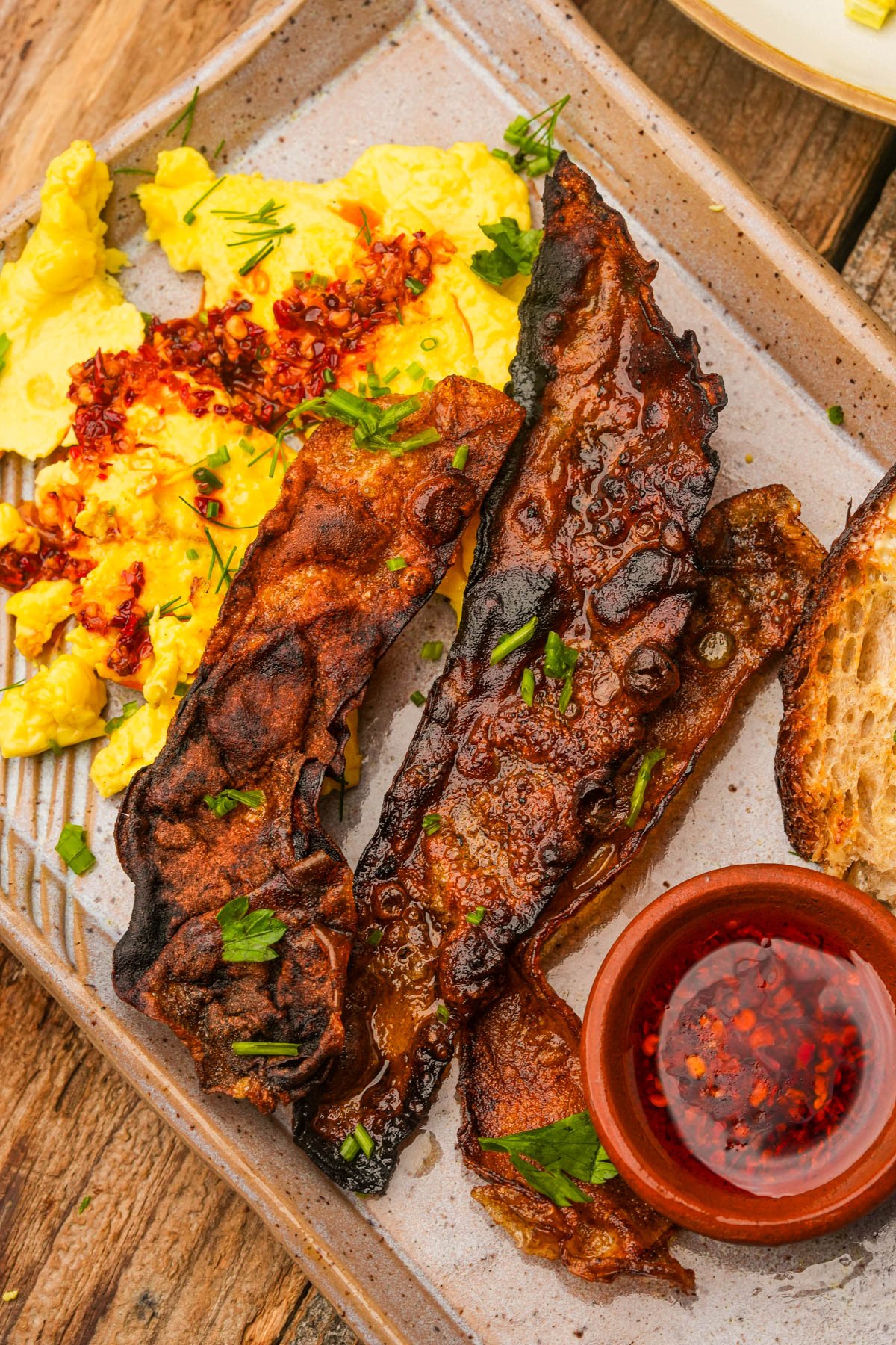 A plate with vegan scrambled eggs topped with chili flakes, three strips of crispy rice paper bacon, a slice of toasted bread, and a small bowl of chili oil on a wooden table.