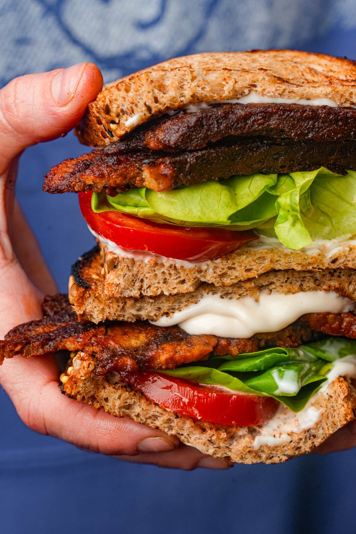 A close-up of a hand holding a sandwich with whole wheat bread, lettuce, tomato, mayonnaise, and crispy dark strips of seitan bacon.