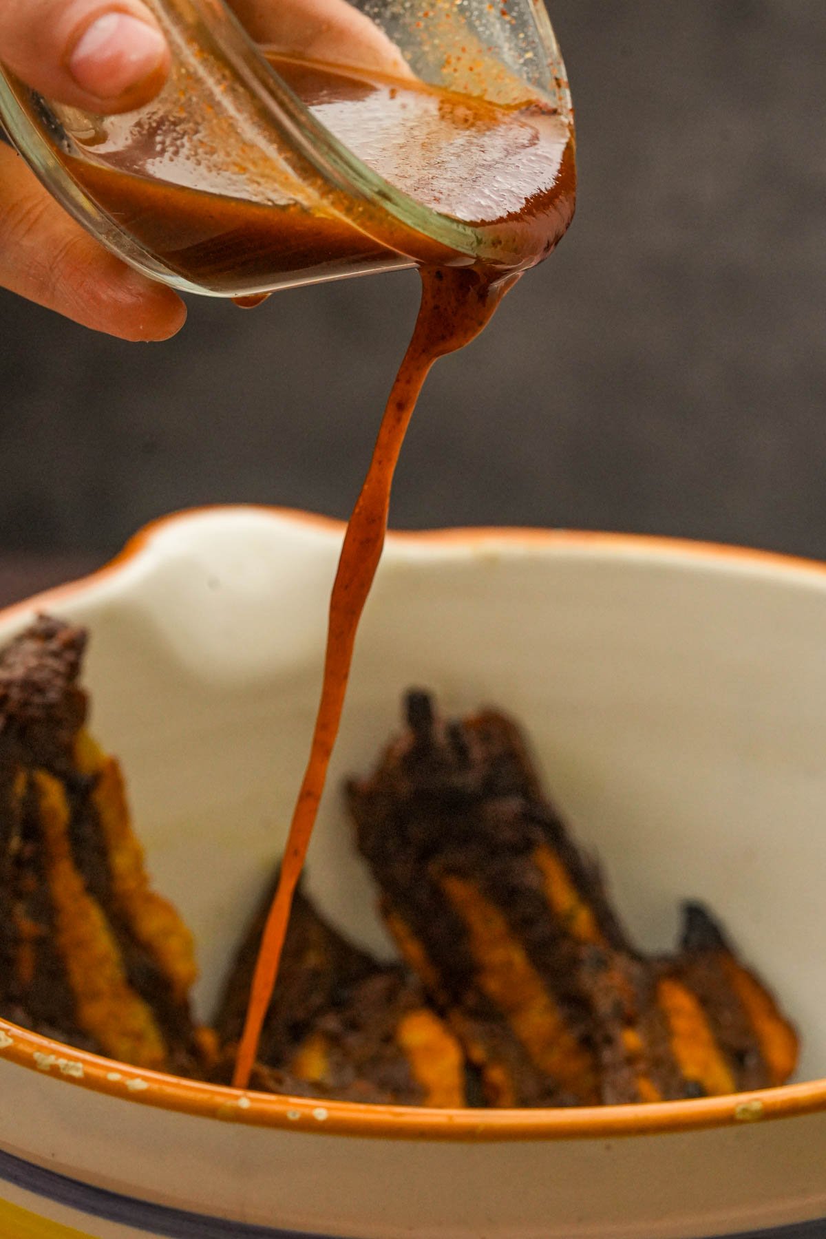 A hand pours vegan bacon marinade from a glass container into a bowl containing strips of seitan bacon.