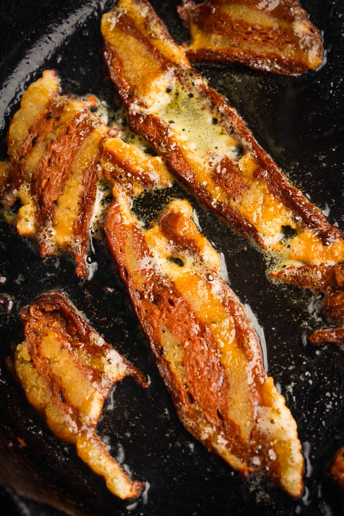 Several strips of seitan bacon are frying in a pan, with visible grease and bubbles around the crispy, browned edges.