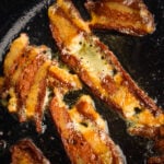 Close-up of several strips of sizzling seitan bacon cooking in a pan, with visible grease and bubbles forming around the edges.