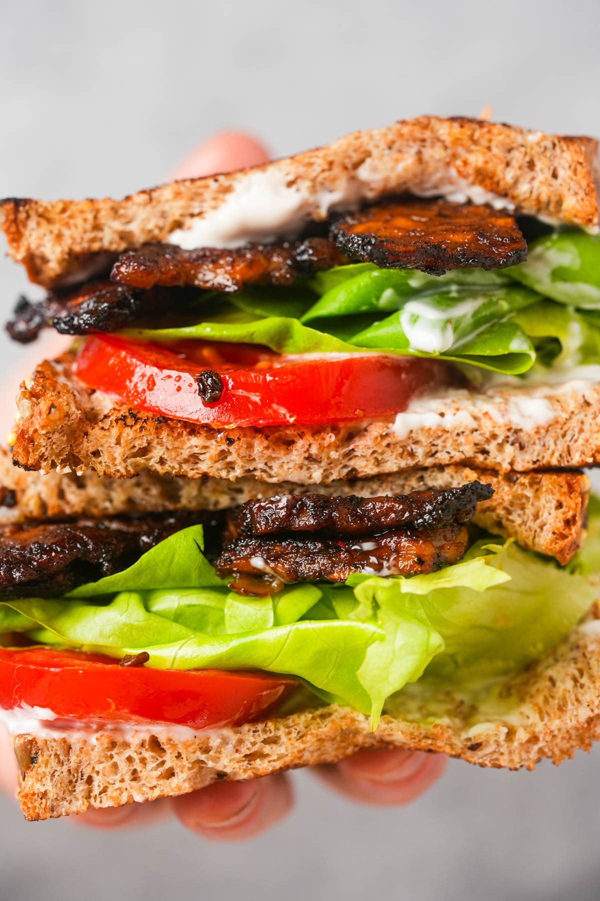 A hand holds a sandwich made with whole grain bread, lettuce, tomato slices, vegan bacon, and mayonnaise.