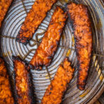 Several strips of cooked tempeh bacon on a round wire rack in a metal tray, with visible caramelization and crispy edges.