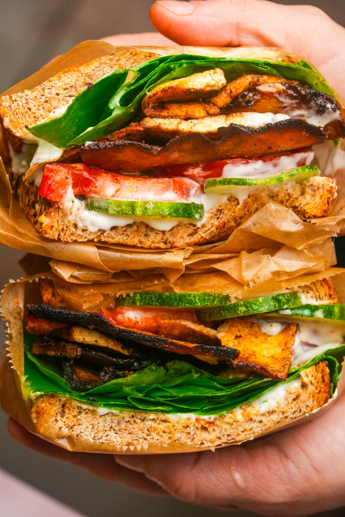 Two halves of a sandwich with lettuce, tomato, cucumber, creamy spread, and charred tofu bacon between whole grain bread, wrapped in brown paper, held in a hand.