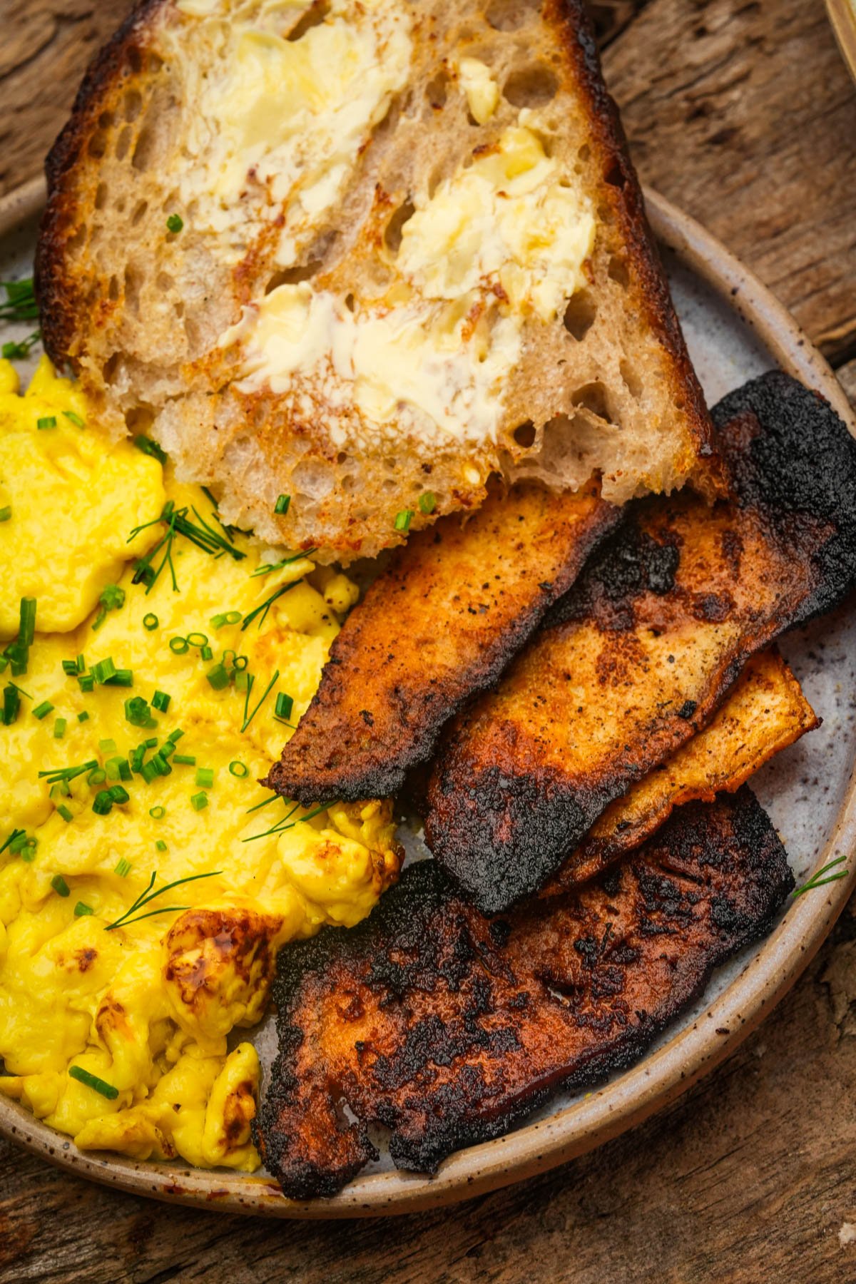 A plate with vegan scrambled eggs topped with chives, three pieces of charred tofu bacon, and a slice of buttered bread on a rustic wooden surface.