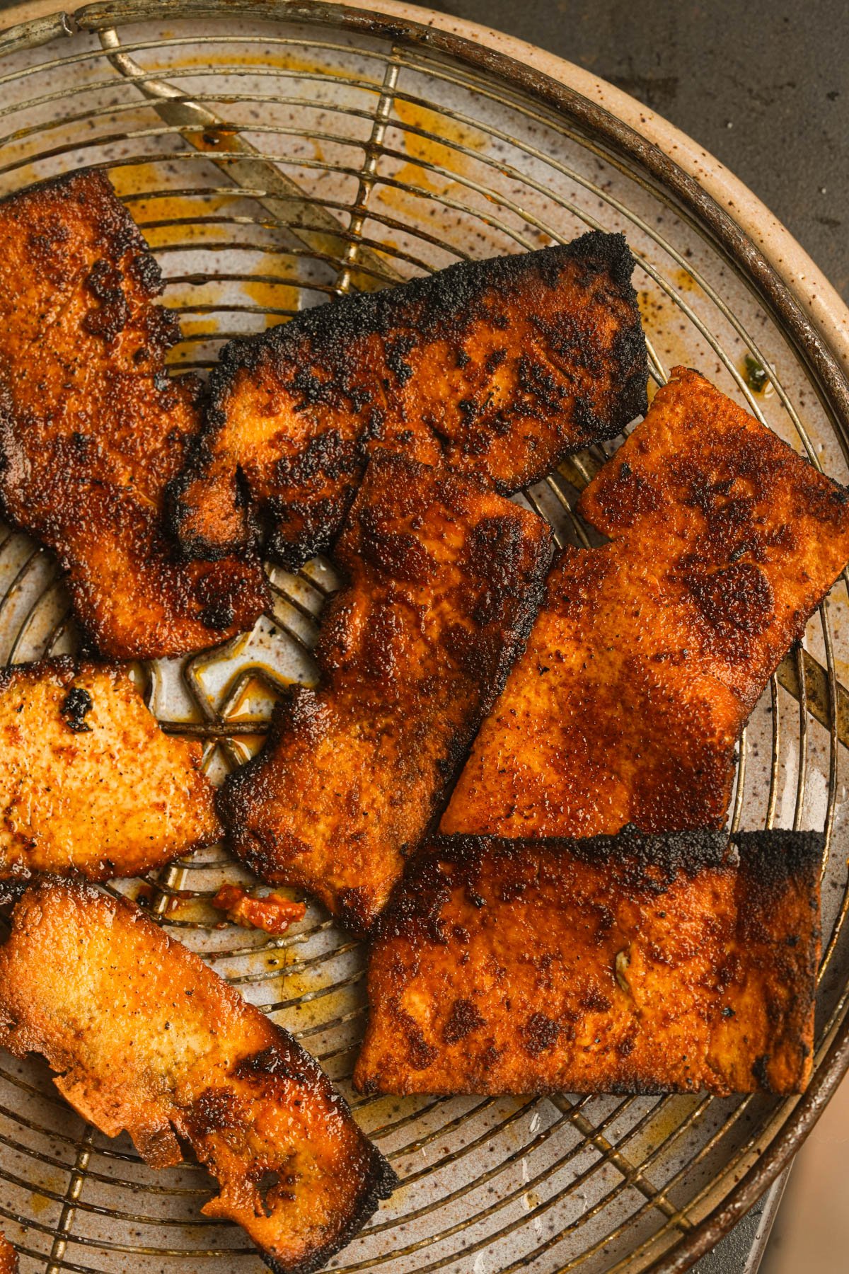 Several pieces of well-seasoned, charred vegan bacon rest on a metal cooling rack with some oil drippings visible.
