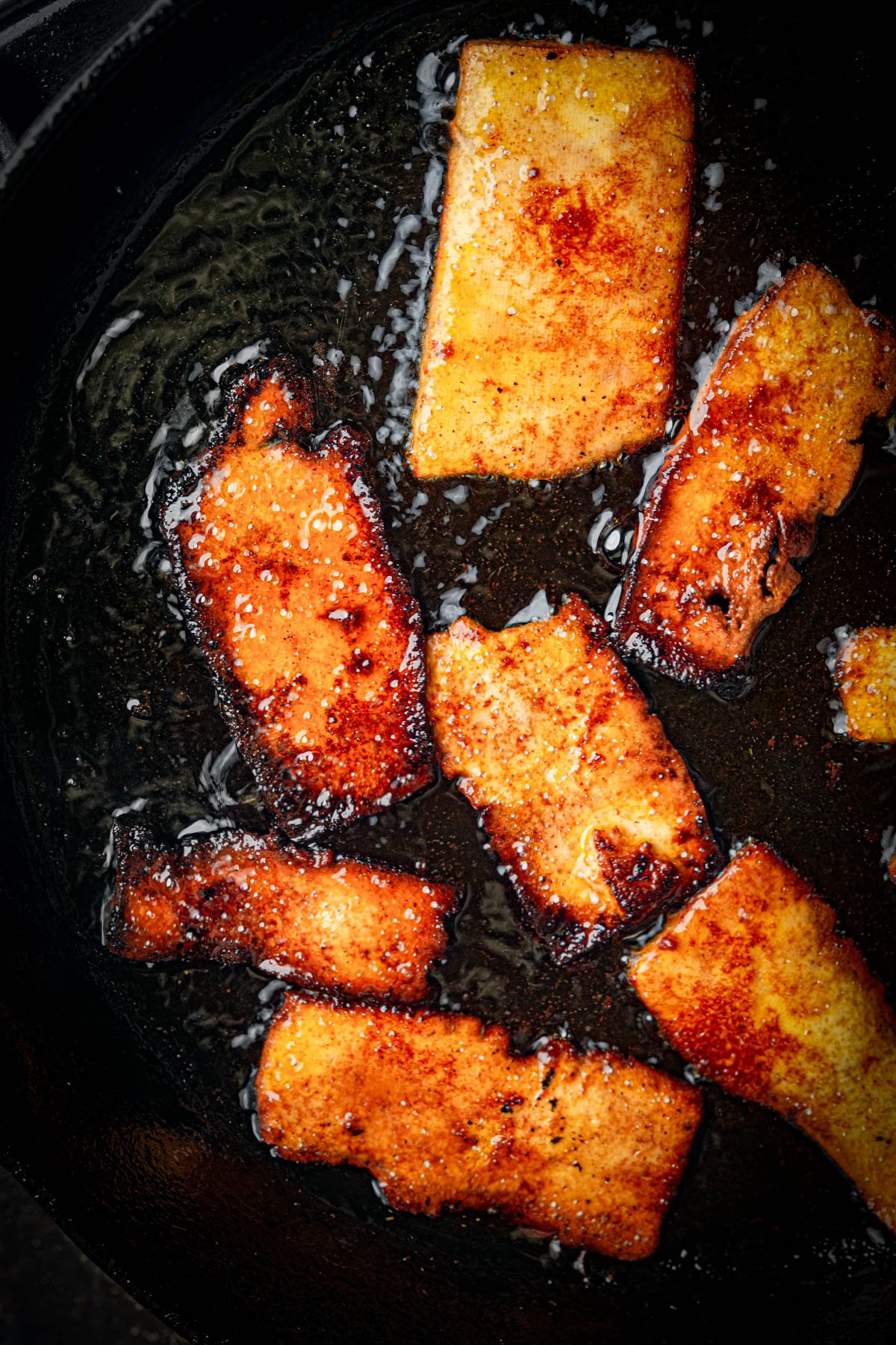 Sliced pieces of tofu frying in oil in a pan, with a golden-brown, crispy, and slightly charred coating.