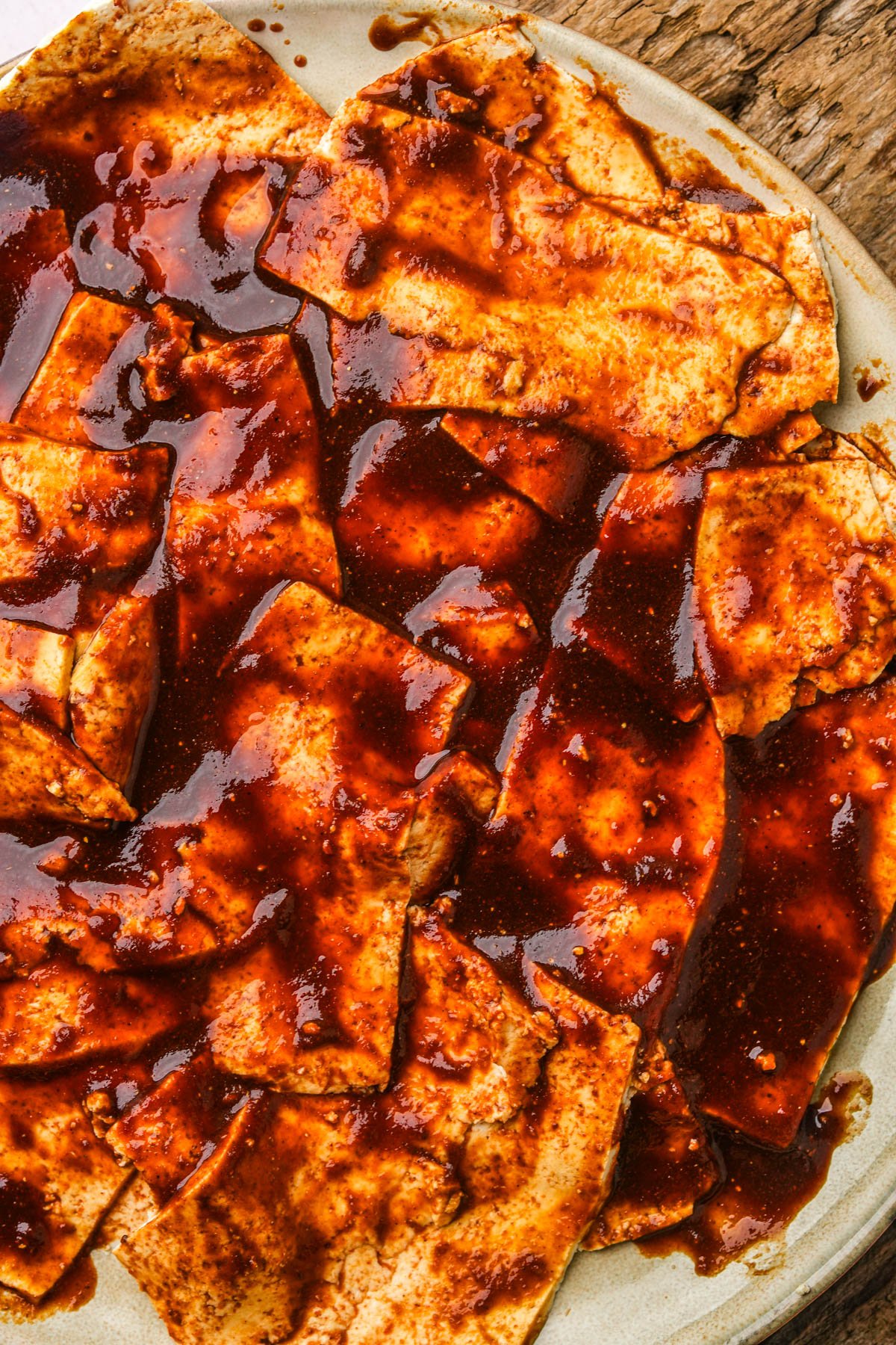 A plate of uncooked tofu bacon covered in a dark marinade on a rustic wooden table.
