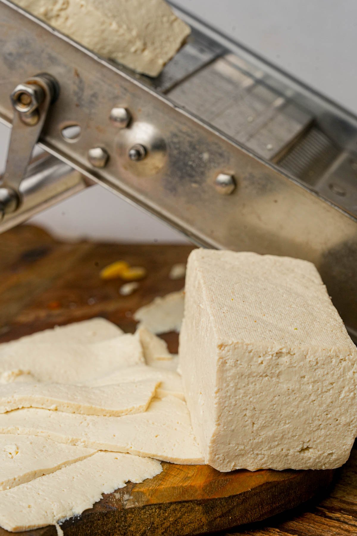 Block of tofu on a wooden board with several thin slices cut, metal mandoline slicer in the background.