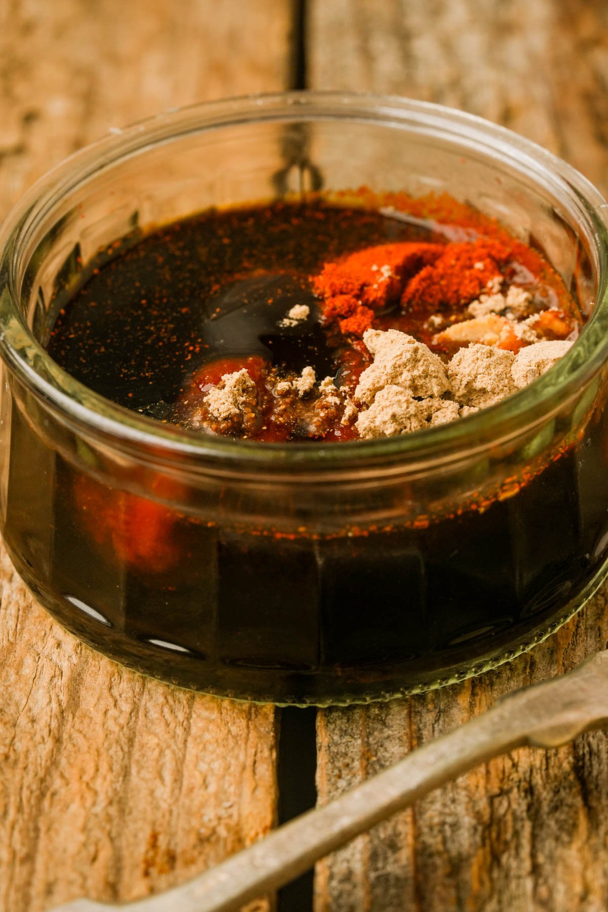 A glass bowl filled with vegan bacon marinade, topped with various powdered spices, sits on a rustic wooden surface.