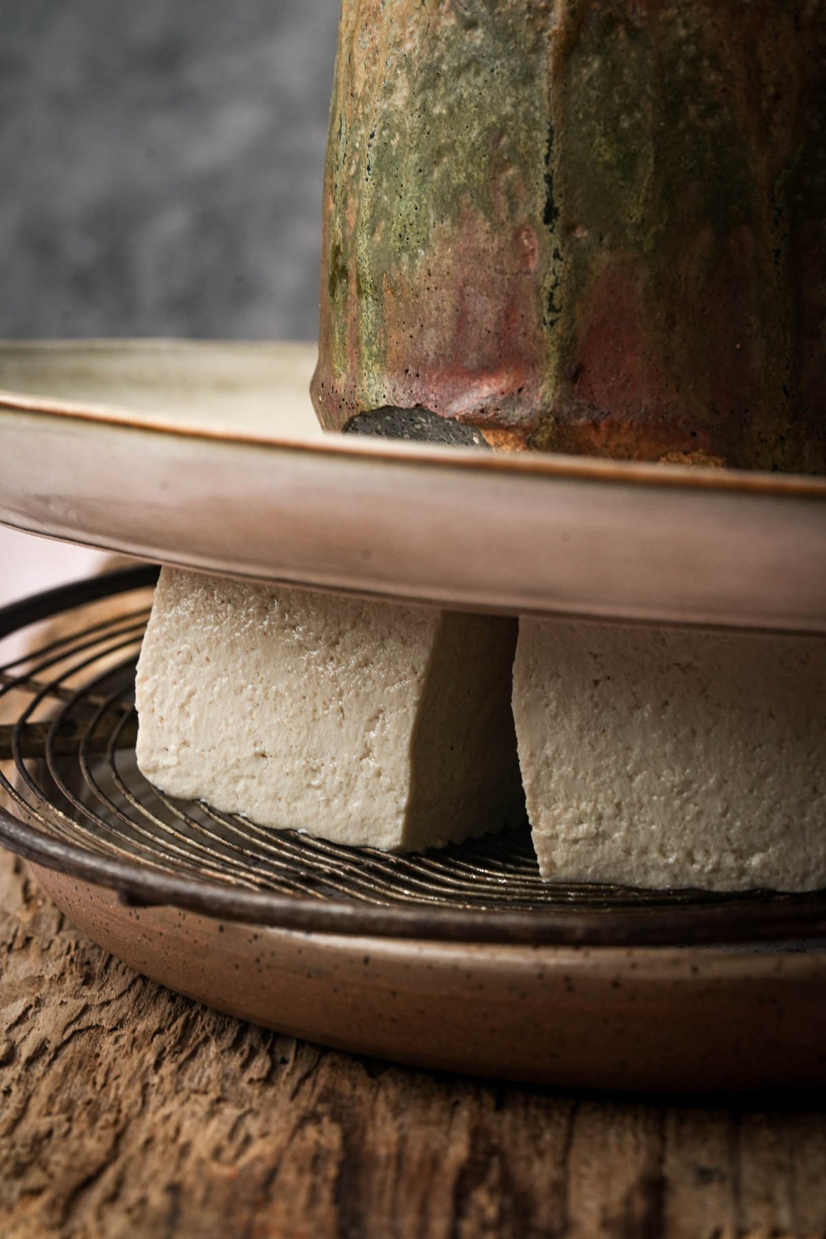 Two blocks of tofu are being pressed under a heavy dish with a wire rack between them, on a rustic wooden surface.