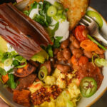 A vegan breakfast bowl with scrambled eggs, pinto beans, tomatoes, hash browns with sour cream and green onions, avocado, sweet potato chips, vegan bacon, salsa, and jalapeño slices.