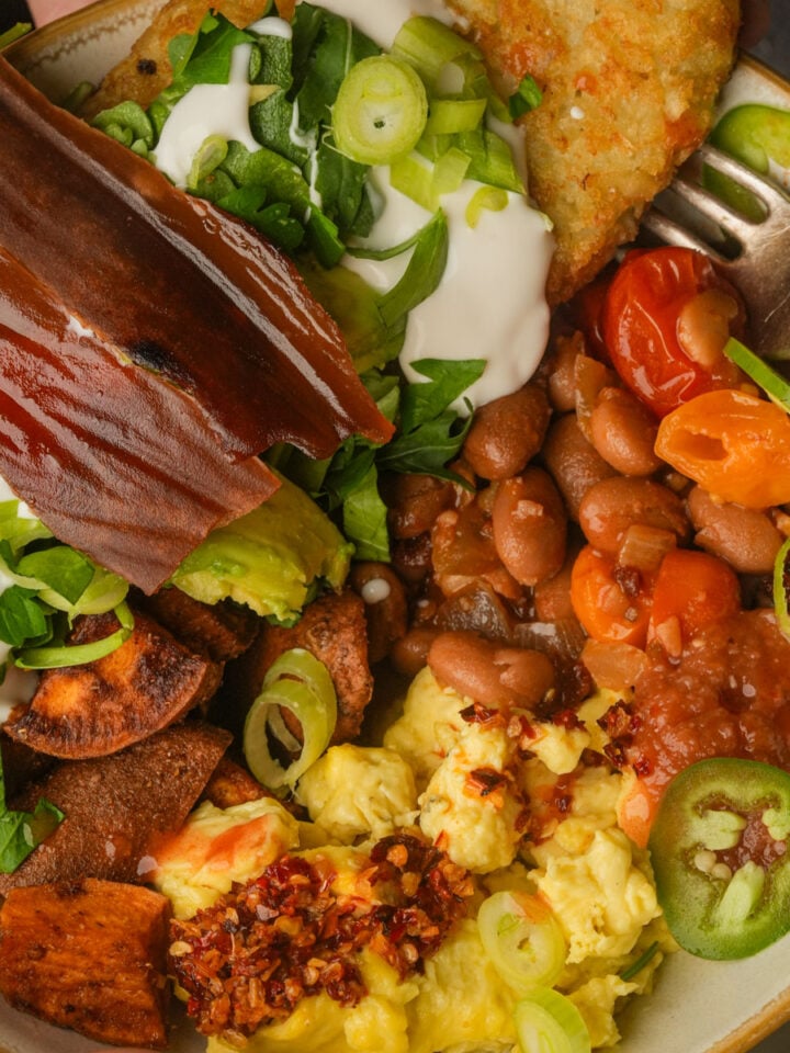 A vegan breakfast bowl with scrambled eggs, pinto beans, tomatoes, hash browns with sour cream and green onions, avocado, sweet potato chips, vegan bacon, salsa, and jalapeño slices.