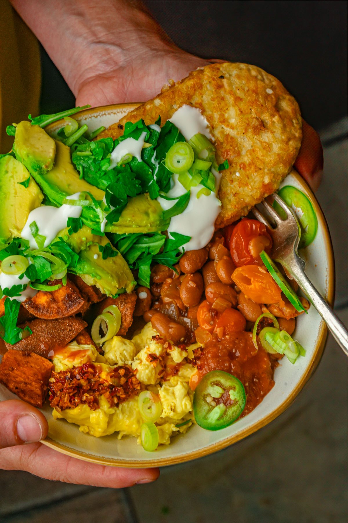 A person holds a vegan breakfast bowl filled with scrambled Just Egg, beans, roasted sweet potatoes, avocado slices, sour cream, green onions, cilantro, a hash brown, salsa, and jalapeño slices.