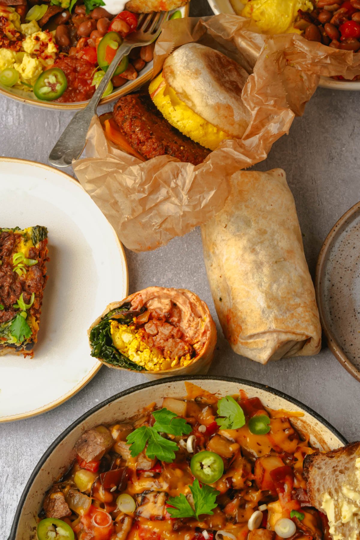 A variety of breakfast foods including a vegan breakfast burrito, vegan breakfast sandwich with plant-based patty, vegan breakfast casserole slice, and vegan breakfast hash on a gray surface.