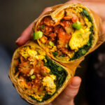 A hand holding a halved vegan breakfast burrito filled with scrambled tofu, greens, sauce, and chipotle grilled seitan.