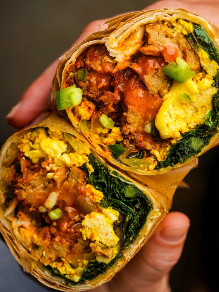 A hand holding a halved vegan breakfast burrito filled with scrambled tofu, greens, sauce, and chipotle grilled seitan.