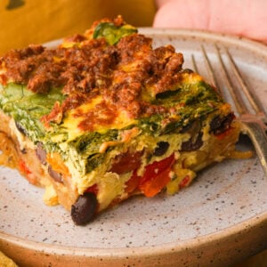 A slice of vegan breakfast casserole topped with spinach sits on a beige plate next to a fork.