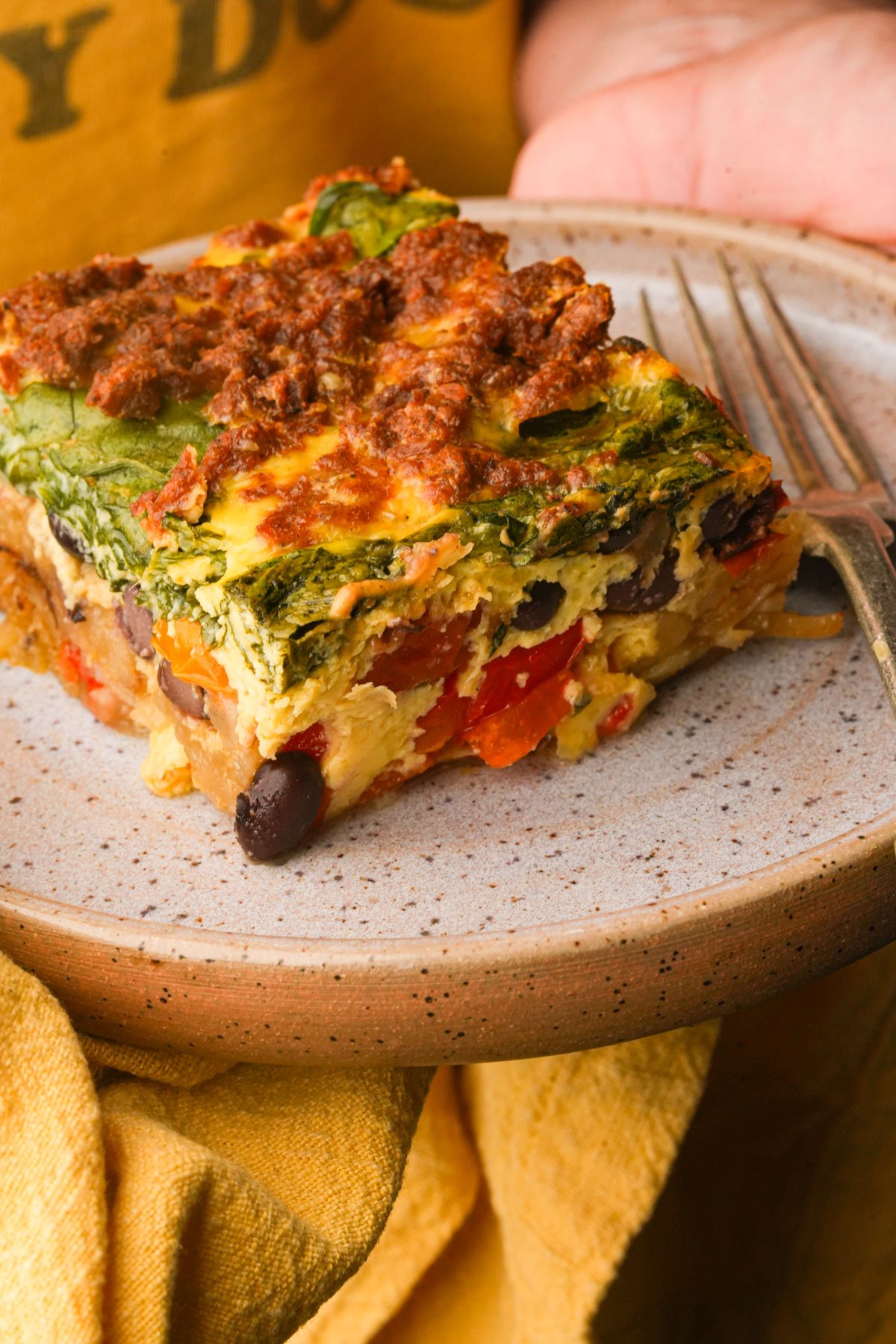 A slice of vegan breakfast casserole with spinach, black beans, and red peppers on a ceramic plate, next to a fork.