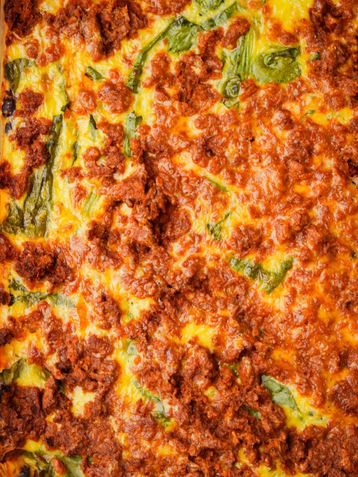A close-up of baked vegan breakfast casserole in a rectangular dish, showing layers of tofu chorizo, spinach, and mung bean egg on top.
