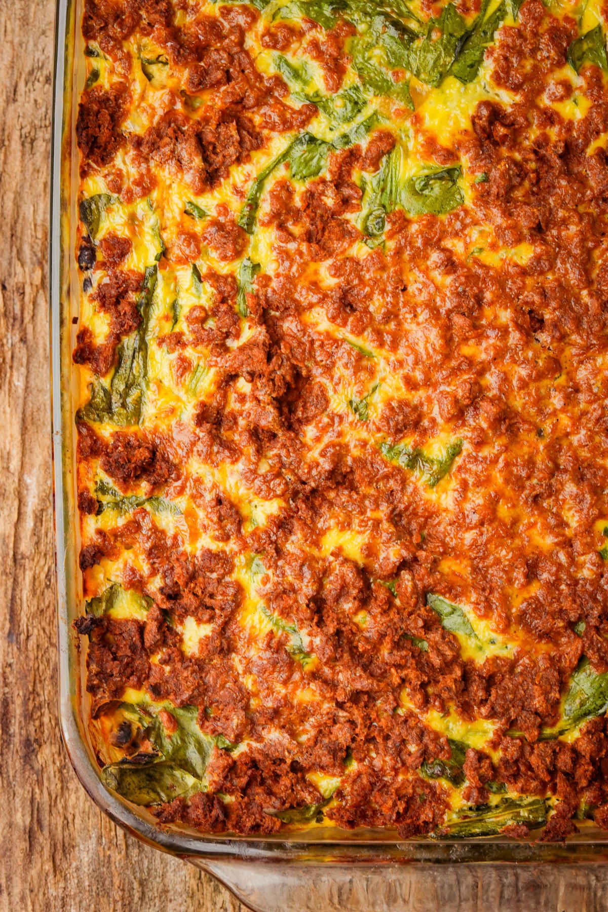 A close-up of a baked vegan breakfast casserole in a glass dish, topped with browned crumbled vegan chorizo, cooked mung bean egg, and visible spinach leaves.