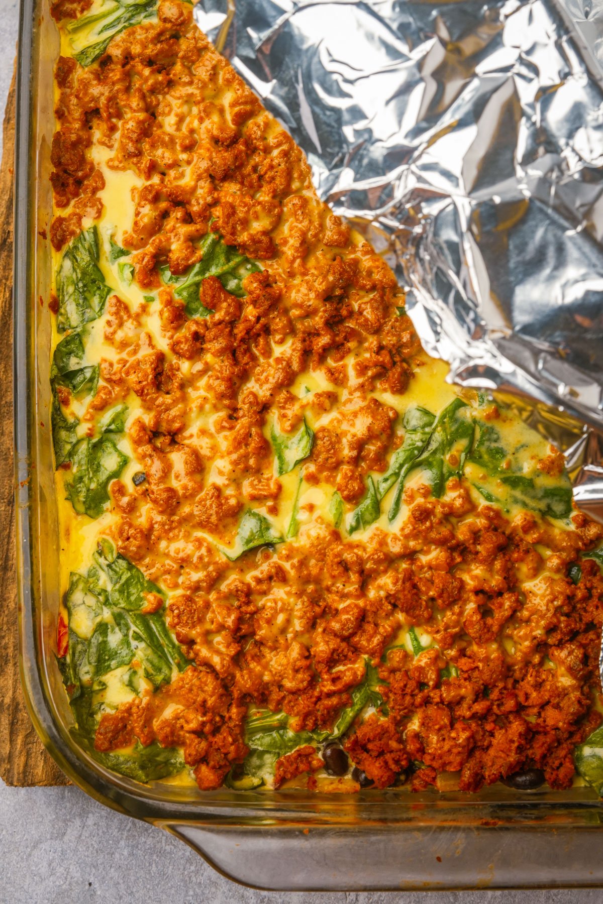 A glass baking dish containing a partially cooked vegan breakfast casserole with spinach and crumbled sausage, next to a sheet of aluminum foil.