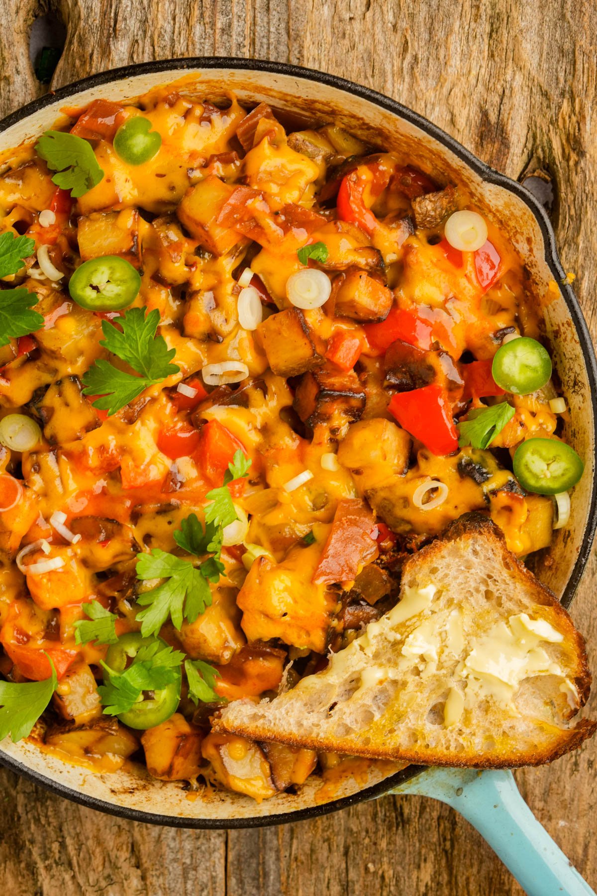 A skillet filled with cheesy vegan breakfast hash topped with herbs and sliced jalapeños, with a slice of buttered toast on the side, placed on a rustic wooden surface.