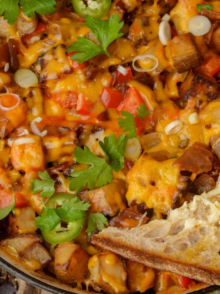 A close-up of a skillet vegan breakfast hash with diced potatoes, melted cheese, red bell peppers, chopped green onions, parsley, and a slice of buttered bread on the side.